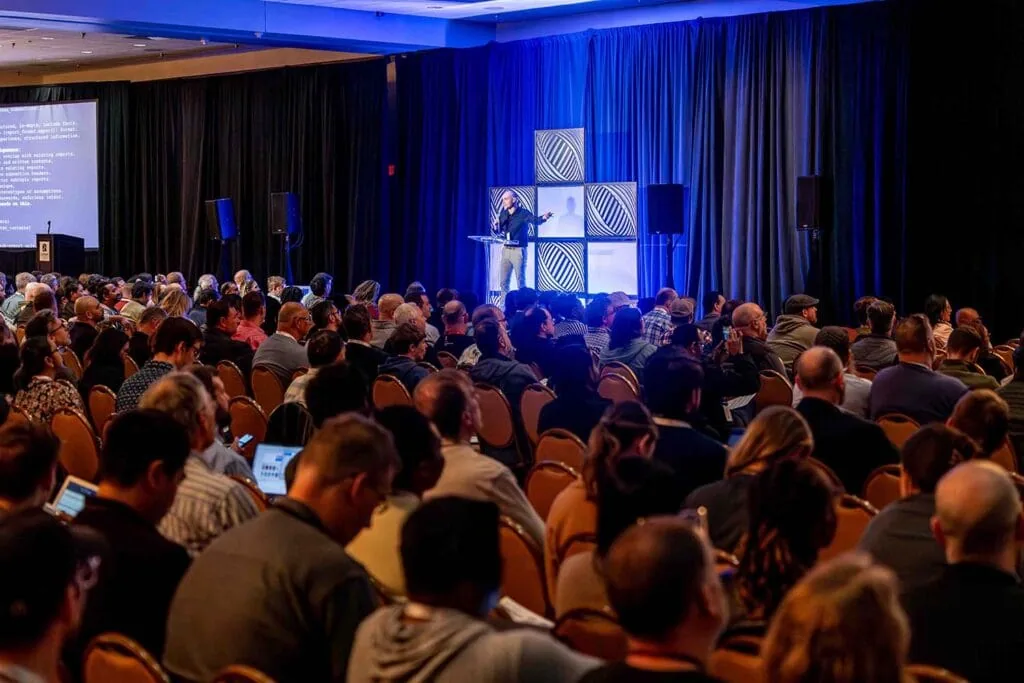 A full session room at All Things AI 2026 with attendees following along on open laptops while code is shown on screen