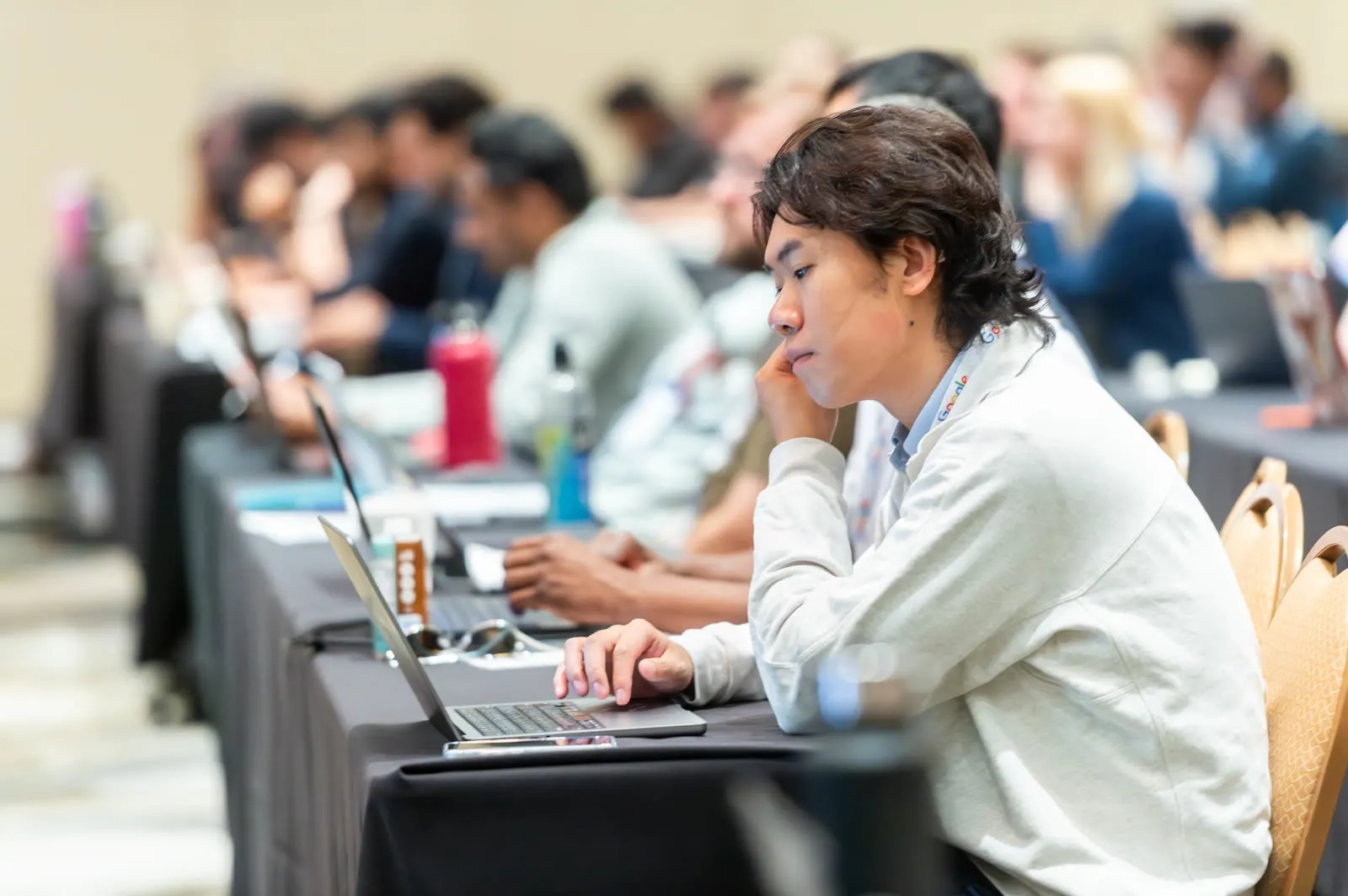 A person concentrating on a laptop at a long workshop table at All Things AI 2026, with a row of others doing the same behind them
