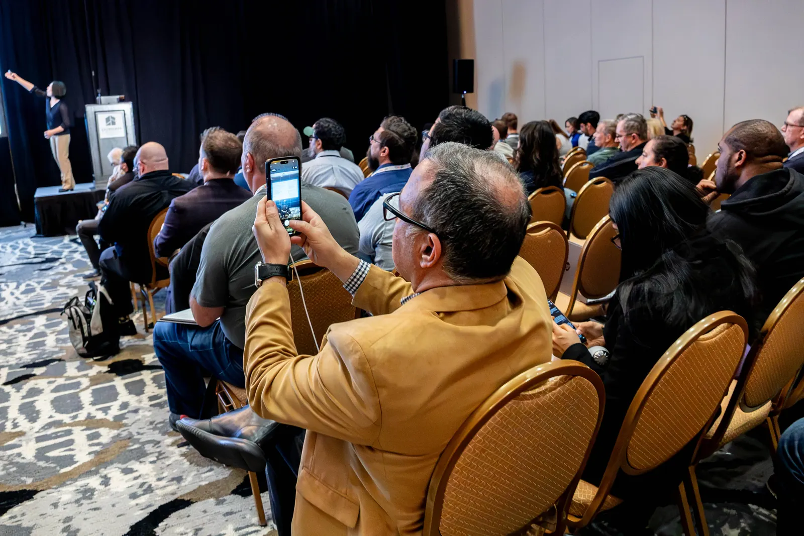 An attendee photographing the presentation while others face the speaker at All Things AI 2026