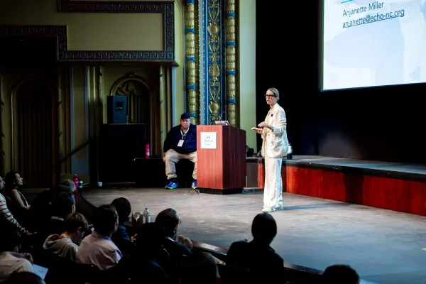 A speaker on the broad Carolina Theatre stage with attendees in the foreground at All Things AI 2026